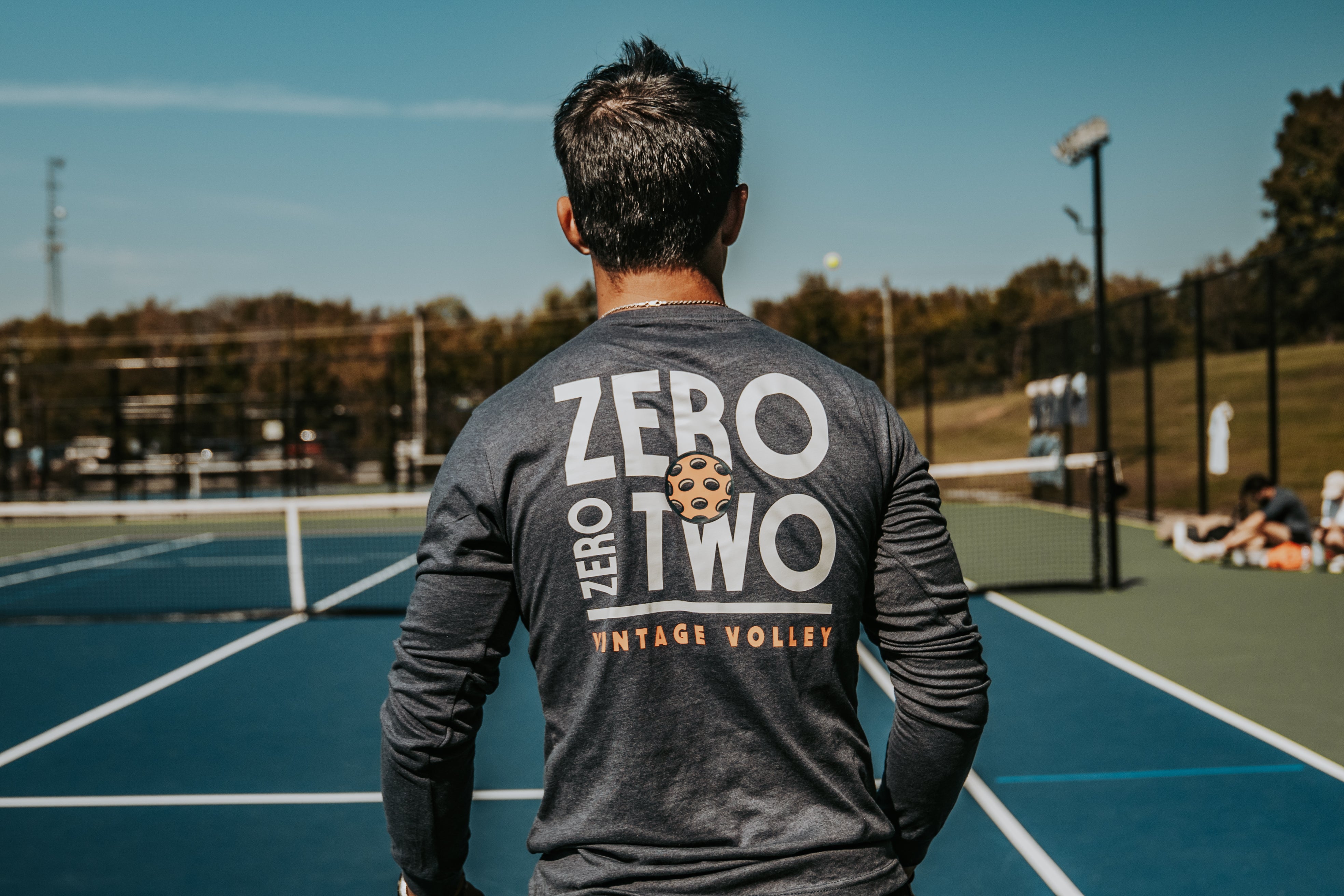 Man wearing a navy blue long-sleeve pickleball shirt with 'ZERO ZERO TWO' and a pickleball graphic on the back, standing on a blue pickleball court under a clear sky, with trees and distant players in the background.
