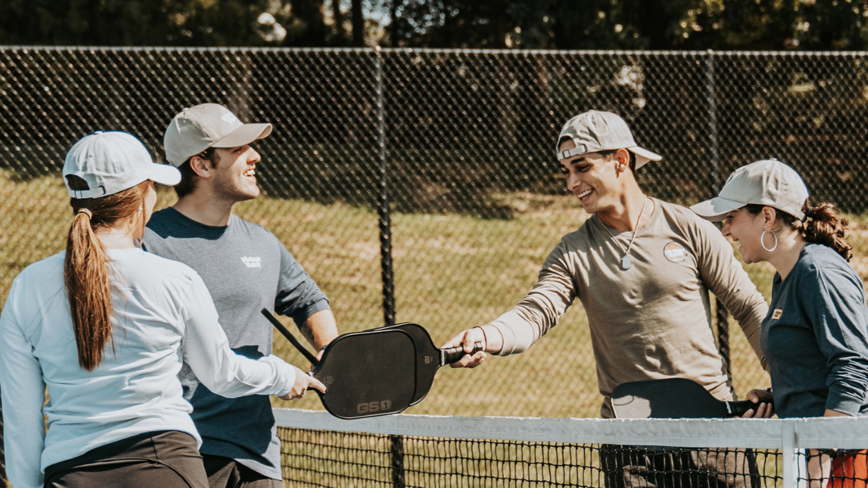 Four individuals on a pickleball court engage in a paddle tap near the net. On the left, a woman with long brown hair tied back and wearing a light blue long-sleeve shirt and black shorts faces away from the camera. Next to her, a man in a light gray cap and navy blue long-sleeve shirt smiles as he extends his paddle forward. Across the net, a young man wearing a backward beige cap, a beige long-sleeve shirt, and a necklace beams as he reaches his paddle forward. To his right, a woman in a gray cap and nav
