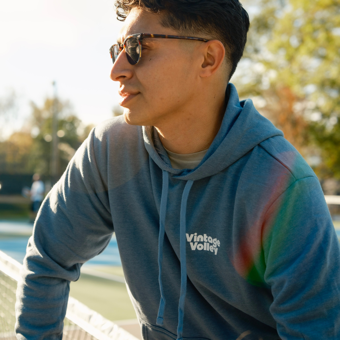 A man wearing a blue hoodie with the "Vintage Volley" logo leans casually against a tennis net on an outdoor court. He gazes to the side with a calm expression. The scene is warmly lit with soft sunlight, and a faint rainbow lens flare appears on the hoodie. The blurred background shows other players on the court, highlighting an active yet relaxed atmosphere.