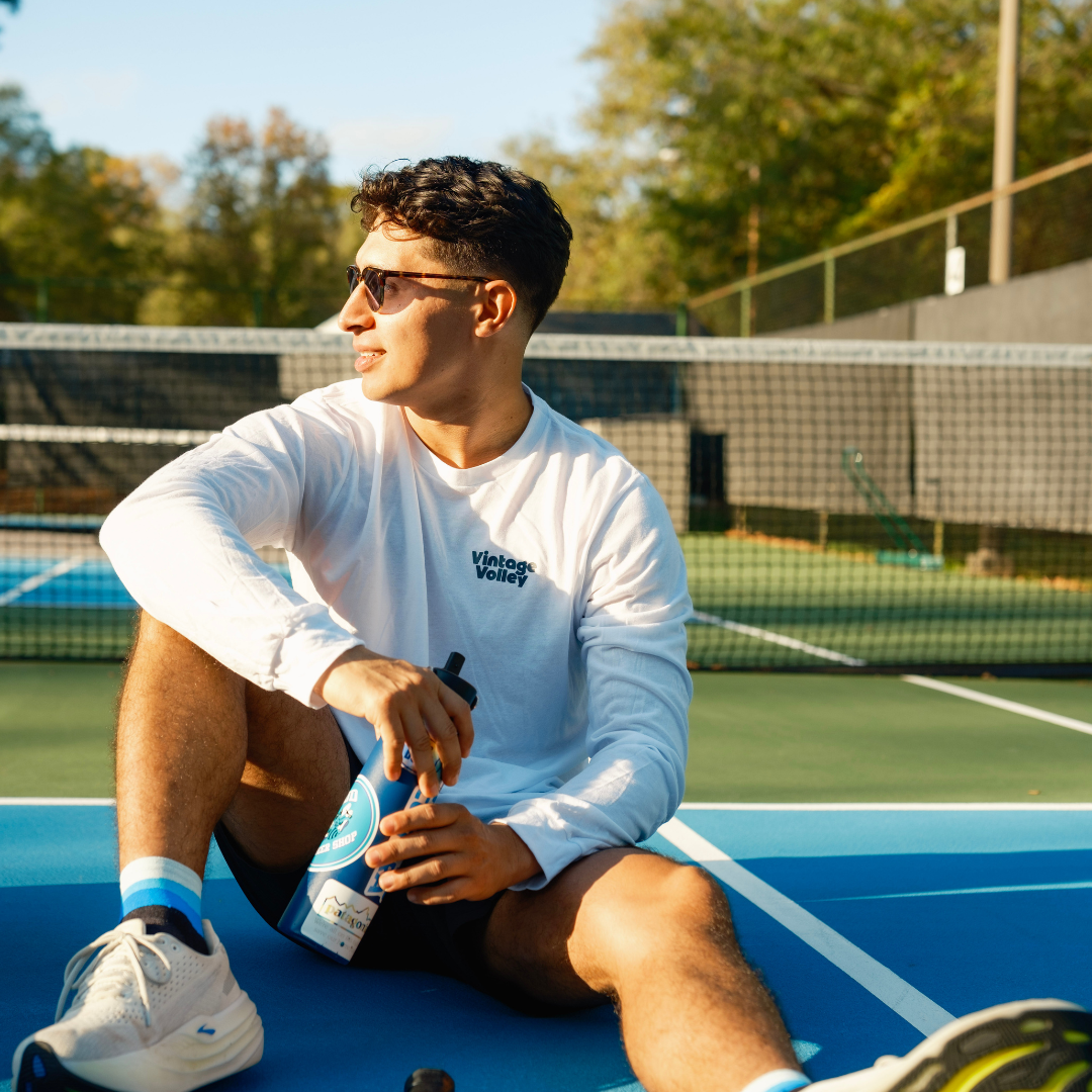 A young man sitting on a pickleball court in a white long-sleeve Vintage Volley shirt, holding a water bottle, with a paddle nearby.