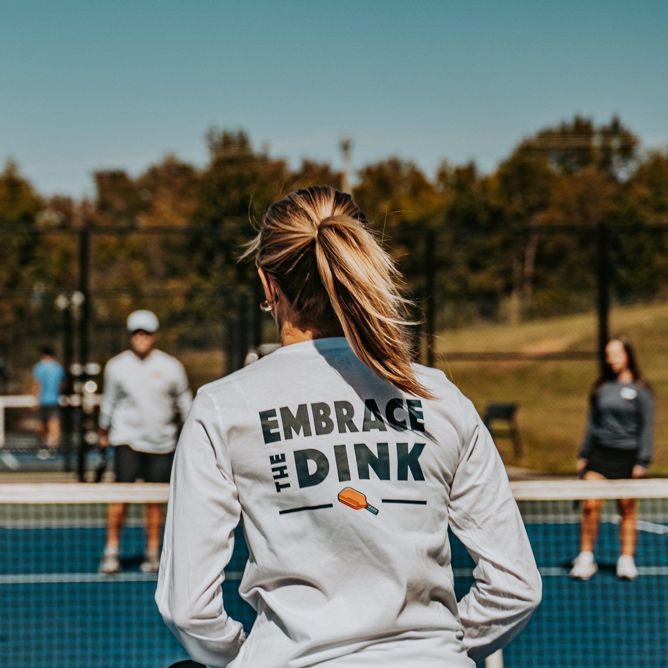 A female pickleball player stands at the net during a match, wearing the Embrace the Dink long-sleeve shirt in White. The back design features bold typography and a colorful paddle graphic, making this lightweight shirt a stylish addition to any pickleball outfit. Perfect for women who love the game and want versatile clothing for on and off the court.