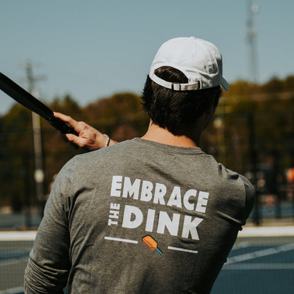 A man is playing pickleball, captured from behind. He is wearing a long-sleeved, sand colored shirt with the phrase "EMBRACE THE DINK" written on the back in bold white text. Below the text is a small image of a pickleball paddle and ball. The man is holding a pickleball paddle mid-swing, and he is wearing a white cap. The background shows an outdoor pickleball court with a few other people visible in the distance, blurred out of focus.