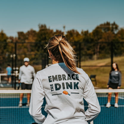 A female pickleball player stands at the net during a match, wearing the Embrace the Dink long-sleeve shirt in White. The back design features bold typography and a colorful paddle graphic, making this lightweight shirt a stylish addition to any pickleball outfit. Perfect for women who love the game and want versatile clothing for on and off the court.