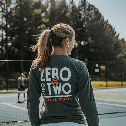 A female pickleball player wearing the Zero Zero Two Vintage Volley Long Sleeve Pickleball Shirt in Gray, standing on a sunny pickleball court. The shirt features bold white text, an orange pickleball graphic, and the words “Vintage Volley.” Designed for lightweight comfort and style, it’s perfect for pickleball games and casual wear, making it a standout piece of pickleball clothing for women.